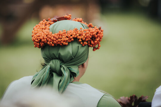 Woman with green headscarf adorned with orange berries sits outdoors, back view, surrounded by blurred greenery and wooden structures in a natural setting