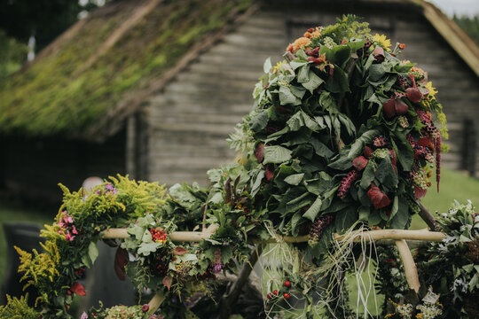 Decorative floral wreath made of green leaves and colorful flowers displayed outdoors near a traditional wooden building with a thatched roof in a rural setting