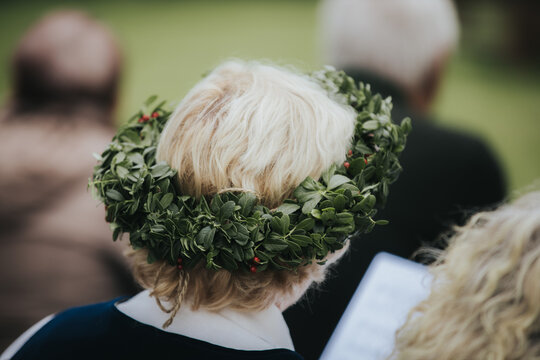 Woman with blonde hair wearing a green floral crown made of leaves and berries, seated outdoors with blurred figures in the background during a celebration or gathering