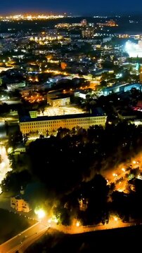 Vertical video. Ryazan, Russia. Aerial night panorama over the illuminated cityscape, the Kremlin Bell Tower's gilded spire rising above historic ramparts as traffic traces golden arteries below. Dro