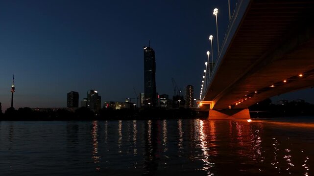 The glowing bridge and modern Vienna skyline reflect gracefully on the dark Danube river at night. Vibrant urban landscapes inspire dynamic economic growth and celebrate advanced city infrastructure.