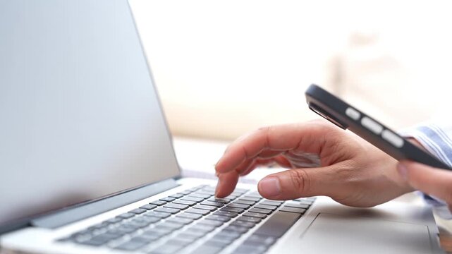 Business employee typing on laptop keyboard while holding smartphone, digital technology workplace with multitasking, online work, professional communication, productivity and modern office workflow.