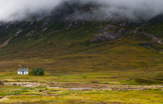Small white cottage beside trees and a stream below grassy rocky mountain slopes partly covered by low cloud. Glencoe Highlands Scotland