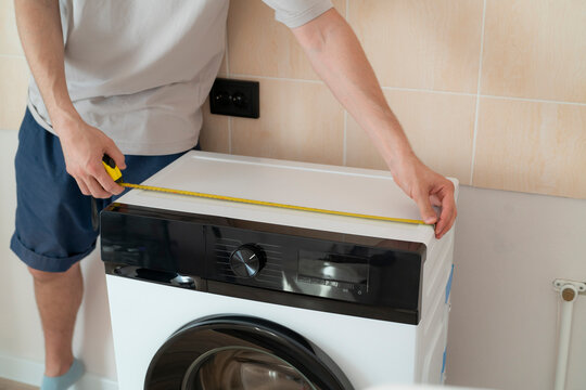 A person stands and uses a tape measure to check the size of a washing machine for fitting in a laundry space
