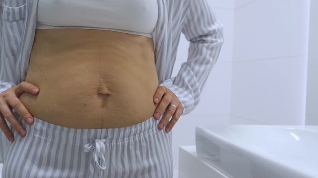 Young adult woman rubs midsection with steady hands, calm in clean bathroom and soft light. Adult woman in striped pajama bottoms and open shirt performs self massage across bare abdomen.