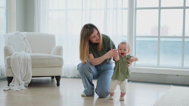 Baby development. First steps. Little infant girl making first steps at home. Happy mother supporting baby first steps. Mom helping toddler child walking in living room. Happy family at home