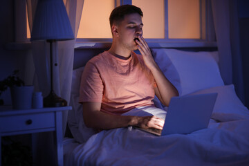 Handsome young man with laptop yawning while sitting in bed at night