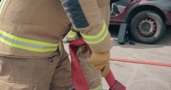 Firefighter team - fireman equipped with emergency services response firefighting crew uniform and gear reeling in fire hose. Department crew from fire station and car wreckage is in scene background.