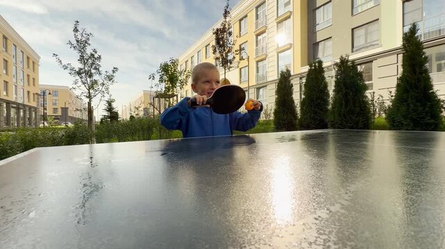 A curious young boy holds a table tennis paddle up in front of his face. He plays ping pong in a sunny courtyard near modern buildings. His playful expression shows pure joy and delight.