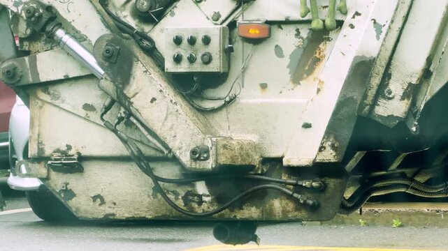 A heavy garbage truck operates its dirty hydraulic mechanism along a city street. The worn metal parts show decades of hard work. The busy industrial scene feels gritty and tired.