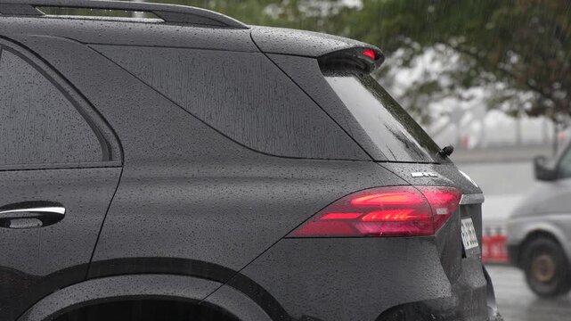 SUV rear quarter with an illuminated tail lamp and rain covered windows as vehicles pass, illustrating automotive signaling and passenger transport on a wet road