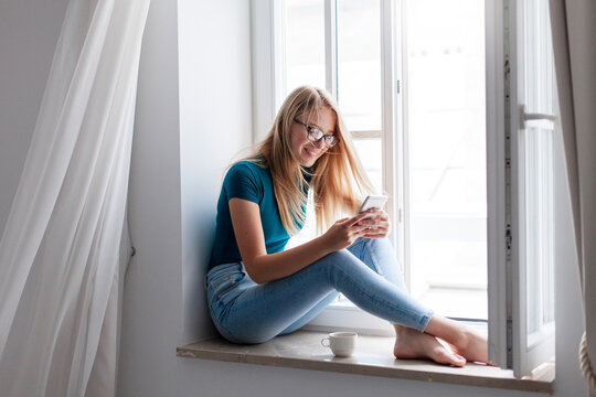 Young woman using smartphone at home. Relaxation with morning coffee on cozy windowsill. Happy girl sitting by opened window, scrolling, browsing. People connection, communication