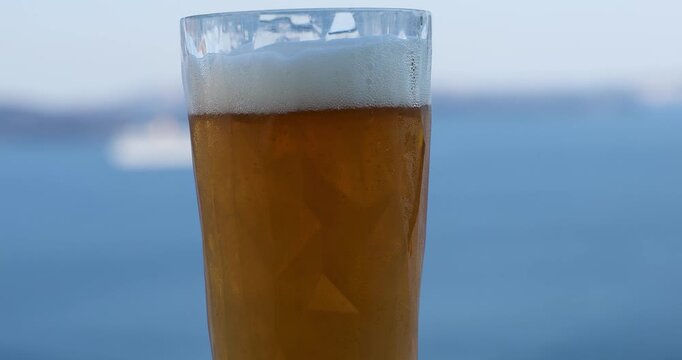 Frothy cold glass of beer on a hot summer day in Greece. Bubbles are rising and the Aegean sea is visible in the background with the Santorini caldera. 