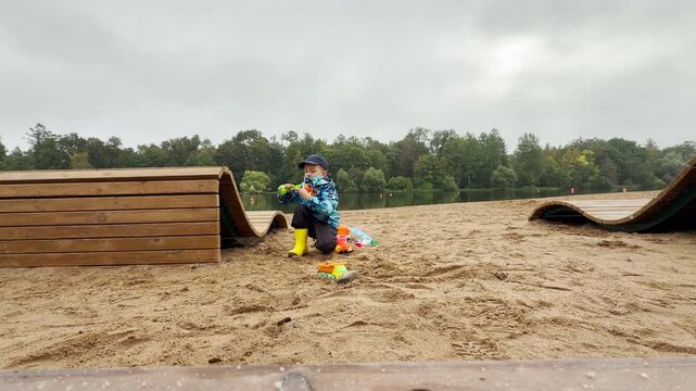 A young boy plays happily with colorful toys on a sandy shore in Pushkin, Saint Petersburg. He sits near wooden benches by a quiet lake. His cheerful focus creates a peaceful mood.