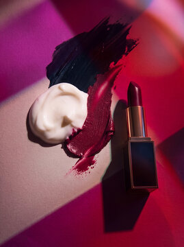 Luxury dark lipstick tube beside swatches of cream, red lipstick, and black texture under vibrant pink geometric light reflections. Abstract beauty concept