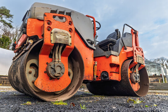 Low-angle ground-level view of heavy tandem drum compactor on freshly laid dark asphalt, hydraulic hoses and hub visible, operator seat and steering wheel behind, trees and sky beyond