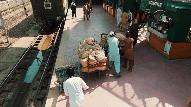 High angle shot of railway porters pushing heavy carts loaded with mail bags along a busy station platform next to train tracks in South Asia.