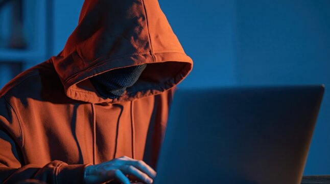Hacker in red hoodie and mask using laptop in dark room