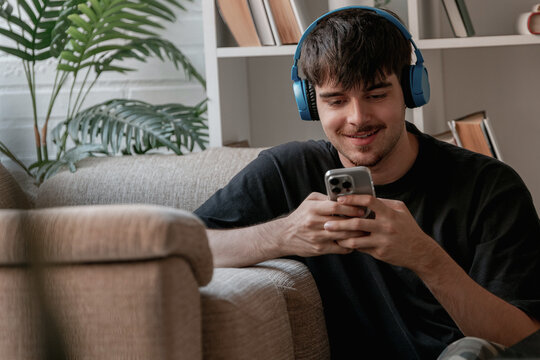 Smiling young Caucasian man wearing blue wireless headphones, relaxing on a sofa at home and using a smartphone, enjoying music and technology in a cozy living room