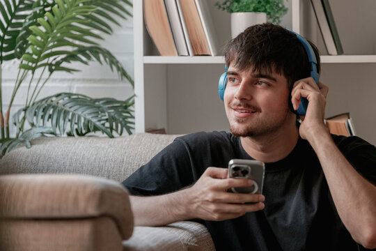 Smiling young Caucasian man wearing blue wireless headphones, relaxing on a sofa at home and using a smartphone, enjoying music and technology in a cozy living room