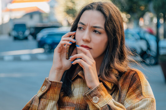 Young woman with worried expression talking on smartphone outdoors, autumn street scene with warm plaid jacket, pensive and concerned mood, urban lifestyle photography