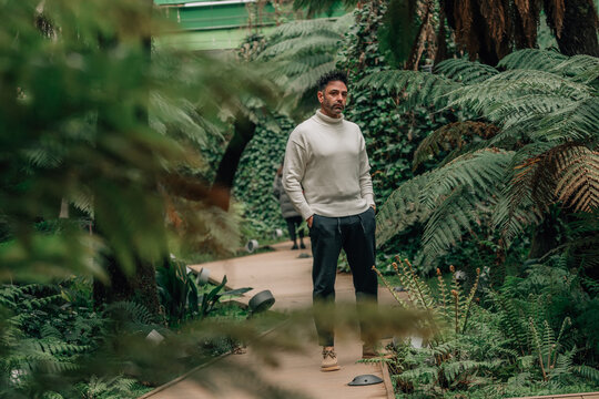 Confident adult man standing on a wooden walkway in a lush botanical garden greenhouse, surrounded by tropical ferns and greenery