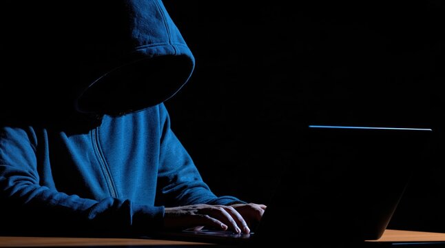 Anonymous hacker in blue hoodie typing on laptop computer in dark environment