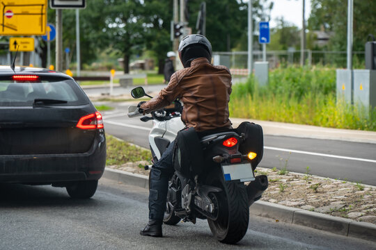 Helmeted motorcyclist with pannier bags stopped beside a car with one foot on the road, illustrating motorcycle commuting and vehicle positioning at a junction