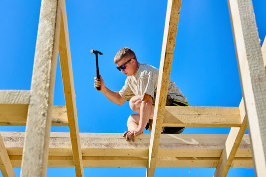 Working at height as young construction worker uses hammer to fasten timber boards on unfinished wooden frame.