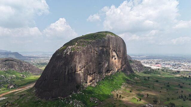 Aerial view of a massive granite rock formation surrounded by green farmland and a sprawling Nigerian landscape.