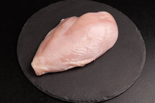 Raw chicken breast background closeup with single fresh poultry fillet chunk on black slate plate, organic meat protein ingredient top view flat lay over dark surface, horizontal minimal copy space.