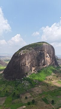 Vertical aerial view of a massive granite rock formation surrounded by green farmland and a sprawling Nigerian landscape