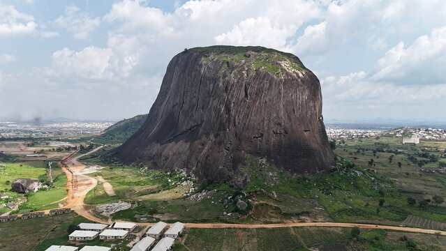 Aerial view of a massive granite rock formation surrounded by green farmland and a sprawling Nigerian landscape.