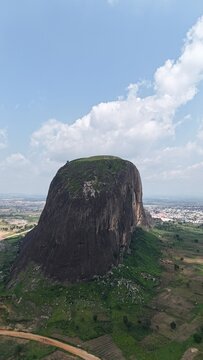 Vertical aerial view of a massive granite rock formation surrounded by green farmland and a sprawling Nigerian landscape