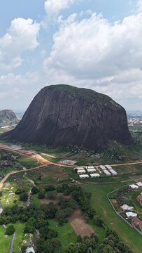 Vertical aerial view of a massive granite rock formation surrounded by green farmland and a sprawling Nigerian landscape