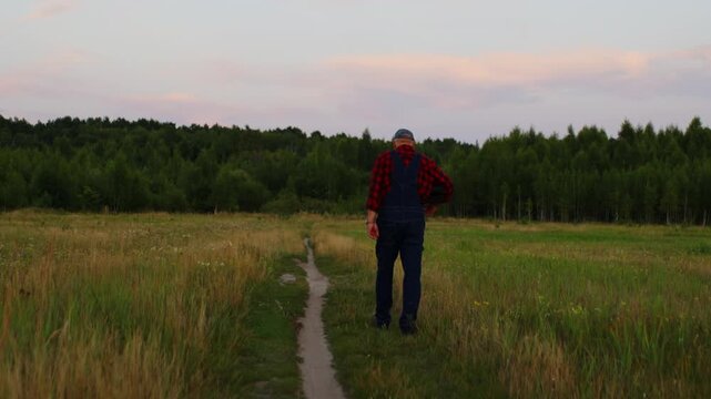 Elderly farmer in plaid shirt and overalls walking alone along narrow dirt path through peaceful green meadow at sunset with forest trees on the horizon