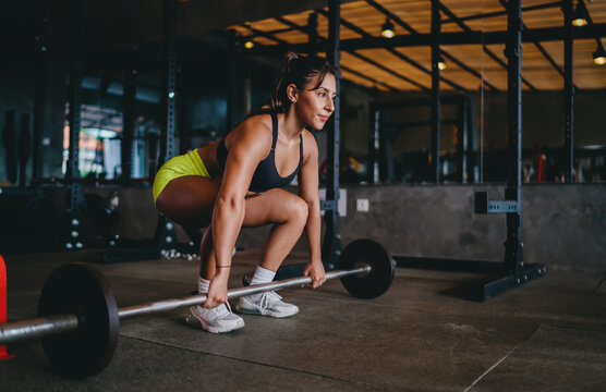 Strength training functional fitness setup allows determined Hispanic female athlete hold weighted barbell near floor inside modern gym room, pursuing personal athletic goals daily