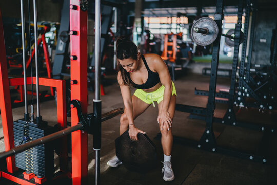 Functional fitness strength training preparation features focused Hispanic female athlete lifting heavy weight plate near barbell machine inside modern gym room, practicing focus daily
