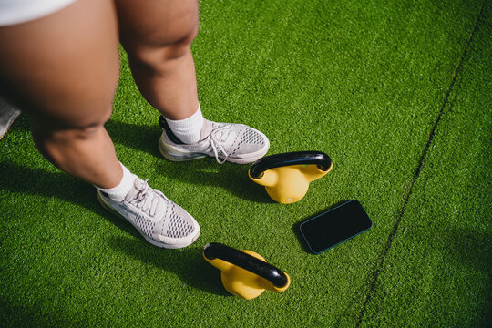 Top view functional fitness perspective features athletic feet standing near yellow kettlebells and smartphone on green artificial turf inside gym room, practicing active wellness goals