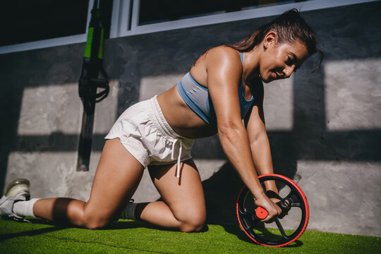 High intensity workout ab roller exercise displays Hispanic female athlete kneeling on artificial grass while holding ab wheel inside modern gym room, practicing active wellness goals