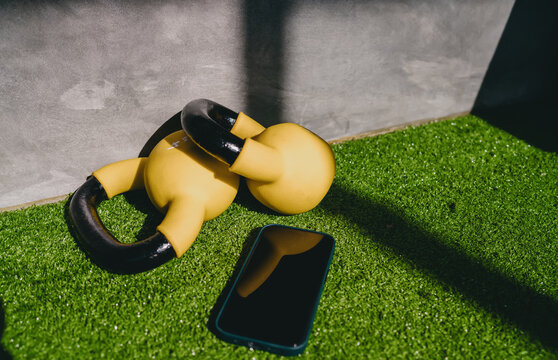 Functional fitness active wellness shot features yellow kettlebells and black smartphone resting on green artificial grass turf inside modern gym room, practicing personal health habits daily