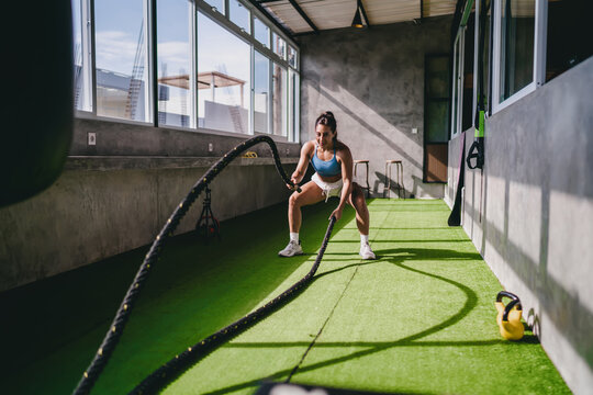 Active wellness high intensity workout routine displays athletic Hispanic female athlete performing battle rope wave exercise on artificial grass inside gym room, pursuing personal fitness goals