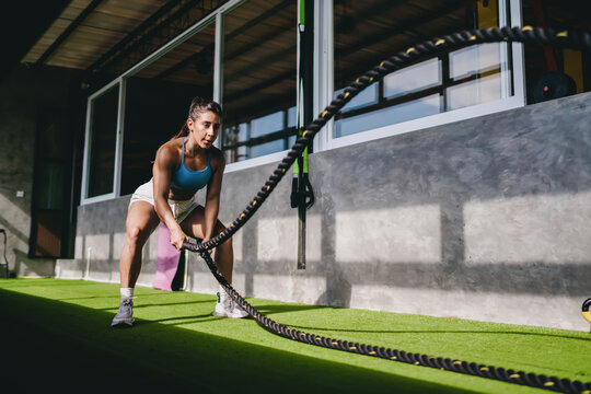 Active wellness functional fitness workout shows athletic Hispanic female athlete battling heavy ropes on artificial grass inside gym corridor, practicing high intensity training goals daily