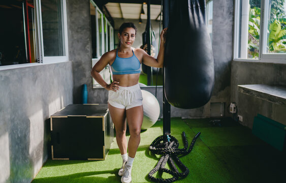 Female empowerment active wellness portrait displays confident Hispanic female athlete standing near heavy punching bag inside gym room, practicing body positivity health goals daily
