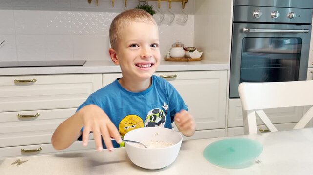 A happy young boy laughs cheerfully with squinted eyes while eating breakfast. He sits at a table in a bright kitchen. His silly expression shows pure morning joy.