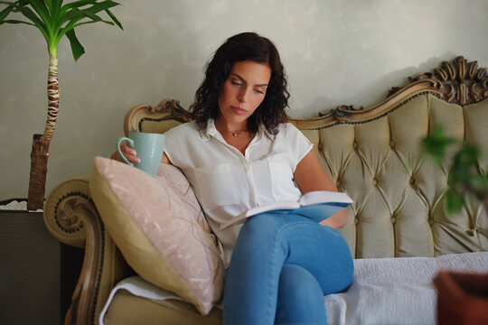 Woman relaxing at home on an antique sofa, engrossed in reading a book, enjoying a warm cup of coffee or tea during her peaceful leisure time indoors