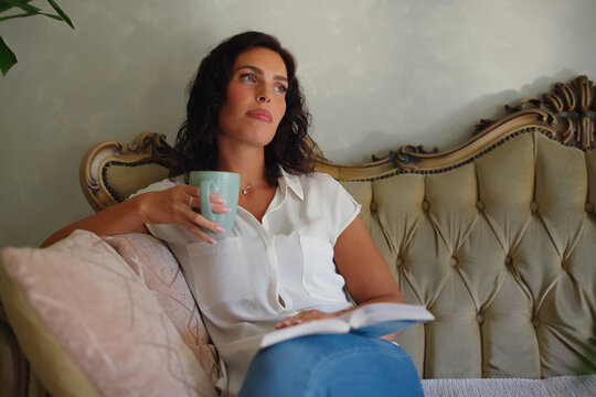 Woman relaxing on a vintage couch, holding a mug and a book, reflecting and contemplating during a quiet moment at home, enjoying peace and solitude