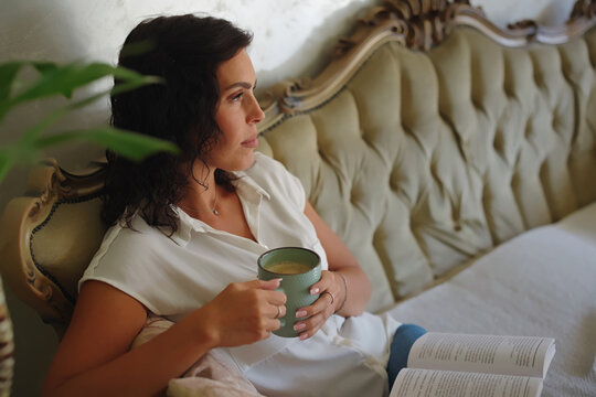 Young woman relaxing on an elegant vintage sofa, enjoying a hot coffee, reading a book, and contemplating with a thoughtful expression in a cozy home environment