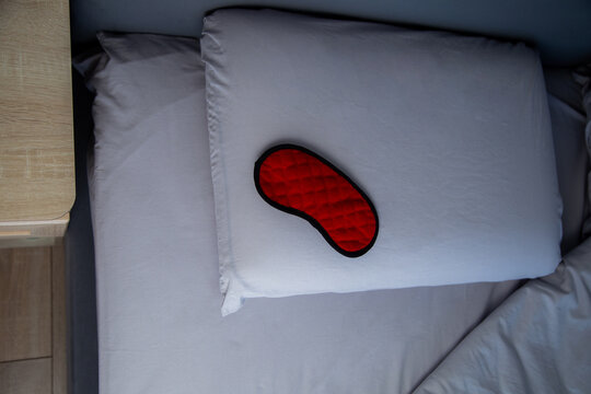 Top view of a red sleep mask lying on a pillow and inviting you to sleep against a background of gray pastel linens and a bedside table. The concept of healthy sleep, sleep patterns, and bedroom