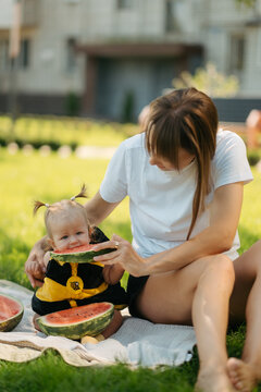 Happy mother and baby girl eating fresh red watermelon slice on green grass outdoors picnic, family summer concept.